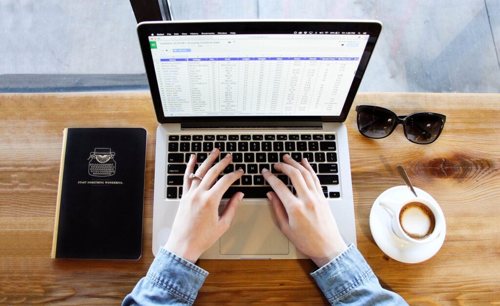 An open notebook laptop showing a spreadsheet being edited. The laptop is rested on a wooden desk with a notepad on the left and sunglasses and a cup of coffee on the right.